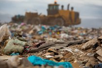 Close up of rubbish with out of focus bulldozer working and clearing rubbish piled on a landfill full of trash with cloudy overcast sky in the background. Global environmental issue of waste disposal. — Stock Photo