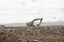 Stormo di uccelli che sorvolano lo scavatore lavorando e ripulendo i rifiuti accumulati su una discarica piena di spazzatura con cielo nuvoloso coperto sullo sfondo. Questione ambientale globale dello smaltimento dei rifiuti. — Foto stock