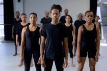 Vista frontale di un ballerino maschio moderno di razza mista che indossa vestiti neri, in piedi di fronte a un gruppo multietnico di ballerini di sesso maschile e femminile in forma, guardando dritto in una fotocamera. — Foto stock