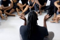 Vista trasera de cerca de un bailarín moderno masculino de raza mixta con traje negro, sentado en el suelo y hablando con un grupo de bailarines durante una clase de baile en un estudio luminoso. - foto de stock
