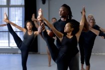 Vue de face d'un danseur moderne masculin mixte en tenue noire, soutenant une danseuse tout en s'étirant lors d'un cours de danse dans un studio lumineux, avec d'autres danseurs exerçant en arrière-plan. — Photo de stock