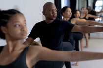Nahaufnahme einer multiethnischen Gruppe fitter moderner Tänzerinnen und Tänzer in schwarzen Outfits, die während eines Tanzkurses in einem hellen Studio an einem Geländer stehen und sich nach oben strecken. — Stockfoto