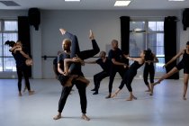 На фоне смешанной расы, современной танцовщицы, одетые в черные костюмы, практикующие танцы во время урока танцев в яркой студии, мужчина держит женщину, позирующую вверх ногами, в то время как другие танцоры стоят на заднем плане. — стоковое фото