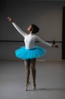 Передній вигляд змішаної раси жіночої балерини в білому трикотажі і синьому турі, танцює в яскравій студії, піднімаючись рукою . — стокове фото