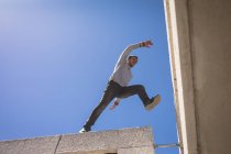 Передній низькокутний вид на кавказького чоловіка, який практикував паркур біля будинку в сонячному дні, стрибаючи на дах.. — стокове фото