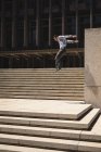 Vista lateral de um homem caucasiano praticando parkour pelo edifício em uma cidade em um dia ensolarado, pulando acima das escadas. — Fotografia de Stock