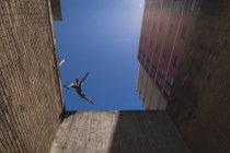 Vista lateral de baixo ângulo de um homem caucasiano praticando parkour pelo prédio em uma cidade em um dia ensolarado, pulando no telhado. — Fotografia de Stock