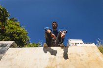 Vorderansicht eines kaukasischen Mannes, der an einem sonnigen Tag in einer Stadt am Gebäude Parkour praktiziert, eine Pause einlegt, sich ausruht und auf einer Mauer sitzt. — Stockfoto