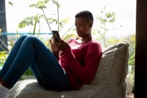 Vue latérale d'une femme afro-américaine assise dans son salon devant une fenêtre par une journée ensoleillée, à l'aide d'un smartphone — Photo de stock