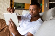 Vista lateral de perto de um homem afro-americano pendurado em sua sala de estar, sentado em um sofá, usando um computador portátil — Fotografia de Stock