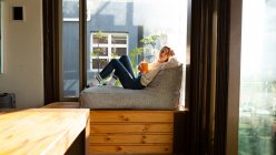 Вид сбоку на кавказку, сидящую в гостиной перед окном в солнечный день, держащую кружку и улыбающуюся — стоковое фото