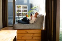 Seitenansicht einer kaukasischen Frau, die an einem sonnigen Tag in ihrem Wohnzimmer vor einem Fenster sitzt, ein Smartphone benutzt, einen Becher hält und lächelt — Stockfoto