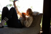 Side view of a Caucasian woman sitting in her living room in front of a window on a sunny day, taking a selfie and holding a mug — Stock Photo