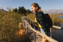 Vista laterale di una donna caucasica che si diverte durante una gita in montagna, in piedi su un ponte, godendo della sua vista, appoggiata su una barriera in una giornata di sole — Foto stock