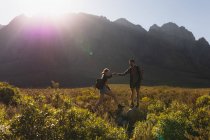 Vista lateral de um casal caucasiano se divertindo em uma viagem para as montanhas, andando em um campo sob as montanhas, um homem está ajudando uma mulher a chegar em uma rocha, em um dia ensolarado — Fotografia de Stock