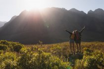 Vorderansicht eines kaukasischen Paares, das eine gute Zeit auf einer Reise in die Berge hat, auf einem Feld unter den Bergen spazieren geht, gemeinsam auf einem Felsen steht, die Hände hebt, an einem sonnigen Tag — Stockfoto