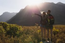 Rückansicht eines kaukasischen Paares, das sich bei einem Ausflug in die Berge amüsiert, auf einem Feld unter den Bergen spaziert, gemeinsam auf einem Felsen steht, eine Frau zeigt auf etwas, an einem sonnigen Tag — Stockfoto