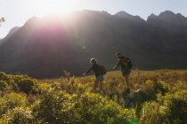 Vista lateral de um casal caucasiano se divertindo em uma viagem às montanhas, andando em um campo sob as montanhas, descendo de uma rocha juntos, em um dia ensolarado — Fotografia de Stock