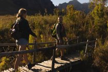 Вид сбоку на кавказскую пару, хорошо проводящую время в поездке в горы, гуляющую по деревянному мосту, смотрящую друг на друга, в солнечный день — стоковое фото