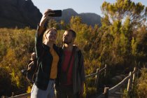 Vue de face gros plan d'un couple caucasien s'amusant lors d'un voyage à la montagne, debout sur un pont en bois, prenant un selfie, par une journée ensoleillée — Photo de stock