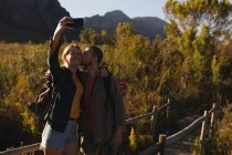 Vista frontal close-up de um casal caucasiano se divertindo em uma viagem para as montanhas, de pé em uma ponte de madeira, tirando uma selfie, um homem está beijando seu parceiro em uma bochecha, em um dia ensolarado — Fotografia de Stock