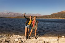 Vue de face gros plan d'un couple caucasien s'amusant lors d'un voyage à la montagne, debout sur un sentier, sur un rivage, prenant un selfie, par une journée ensoleillée — Photo de stock