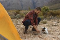 Side view of a Caucasian man having a good time on a trip to the mountains, putting up a tent, with a puppy laying by him — Stock Photo