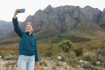 Vista frontal de una mujer caucásica pasando un buen rato en un viaje a las montañas, de pie y tomando una selfie - foto de stock