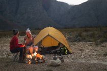 Vista lateral de um casal caucasiano se divertindo em uma viagem às montanhas, sentados ao lado de uma fogueira, conversando, bebendo chá, sorrindo um para o outro — Fotografia de Stock