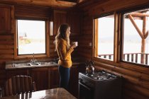 Front view of a Caucasian woman having a good time on a trip to the mountains, standing in a wooden cabin, drinking coffee, looking through the window — Stock Photo