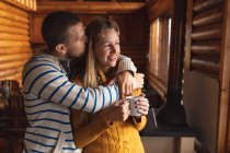 Front view close up of a Caucasian couple having a good time on a trip to the mountains, standing in a cabin, a woman is holding a cup of coffee and a man is kissing her, embracing, looking through  the widow — Stock Photo