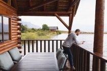 Vista lateral de um homem caucasiano se divertindo em uma viagem às montanhas, de pé em uma varanda em uma cabine, apoiando-se em uma barreira, usando smartphone — Fotografia de Stock