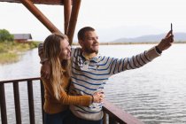 Vista lateral de um casal caucasiano se divertindo em uma viagem às montanhas, de pé em uma varanda em uma cabine, abraçando, tirando uma selfie — Fotografia de Stock