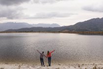 Rear view of a Caucasian couple having a good time on a trip to the mountains, standing on a lake shore, holding hands, raising hands to the air — Stock Photo