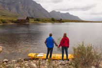 Vista laterale di una coppia caucasica che si diverte durante un viaggio in montagna, si tiene per mano, si gode la vista dopo aver messo un kayak sull'acqua — Foto stock