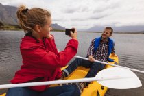 Vista laterale di una coppia caucasica che si diverte durante una gita in montagna, in kayak su un lago, una donna sta fotografando un uomo — Foto stock