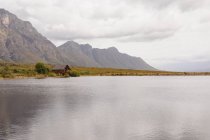 Atemberaubender Blick auf eine Hütte, die einsam an einem See steht, dahinter herrliche Berge und dazwischen ein Feld — Stockfoto