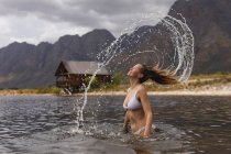 Vista posteriore di una donna caucasica che si diverte durante un viaggio in montagna, in piedi in un lago, gettando i capelli bagnati, lasciando una scia d'acqua in aria — Foto stock