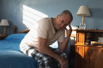 Vista frontal de um homem caucasiano sênior relaxando em casa em seu quarto, sentado ao lado da cama e pensando, olhando para a câmera e sorrindo depois de se levantar de manhã — Fotografia de Stock