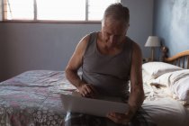 Vista frontale di un anziano caucasico che si rilassa a casa nella sua camera da letto, indossa un giubbotto antiproiettile, si siede sul lato del letto e utilizza il suo computer portatile — Foto stock