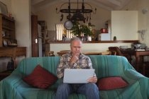 Вид спереди пожилого кавказца, отдыхающего дома в своей гостиной, сидящего на диване с ноутбуком — стоковое фото