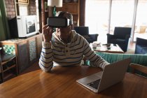 Vorderansicht eines älteren kaukasischen Mannes, der es sich zu Hause in seinem Wohnzimmer gemütlich macht, am Tisch sitzt, einen Laptop benutzt und ein Virtual-Reality-Headset trägt — Stockfoto