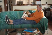 Вид на старшего кавказца, расслабляющегося дома в гостиной, сидящего на диване с поднятыми ногами и играющего со своей домашней собакой — стоковое фото