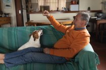 Вид на старшего кавказца, расслабляющегося дома в гостиной, сидящего на диване с поднятыми ногами, играющего со своей собакой и дающего ему угощение — стоковое фото