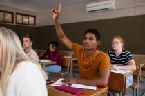Vista frontal de um adolescente mestiço em uma sala de aula sentada na mesa, levantando a mão para responder a uma pergunta, com colegas adolescentes masculinos e femininos sentados em mesas trabalhando em segundo plano — Fotografia de Stock
