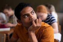 Side view close up of a teenage mixed-race boy in a school classroom sitting at desk, concentrating, with teenage male and female classmates sitting at desks working in the background — Stock Photo