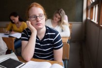 Вид сбоку на девушку-белого подростка в школьном классе, сидящую за столом, сосредоточенную, с подростками-одноклассниками, сидящими за партами, работающими на заднем плане — стоковое фото