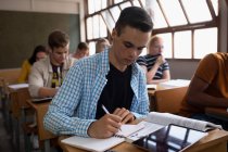 Seitenansicht eines jugendlichen kaukasischen Jungen in einem Klassenzimmer, der konzentriert am Schreibtisch sitzt und schreibt, während im Hintergrund männliche und weibliche Teenager am Schreibtisch sitzen und arbeiten. — Stockfoto