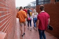 Vista trasera de un grupo multiétnico de alumnos adolescentes de escuelas masculinas y femeninas con bolsos colgando, caminando en sus terrenos de la escuela secundaria, uno con un monopatín - foto de stock