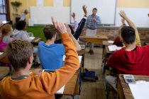 Rückansicht einer multiethnischen Gruppe von High-School-Teenagern in einem Klassenzimmer, die an Schreibtischen sitzen und alle die Hände heben, um eine Frage zu beantworten, während ihr männlicher Lehrer im Hintergrund steht. — Stockfoto